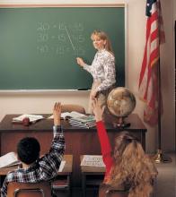 A school teacher is standing in front of a green, framed magnetic chalkboard.